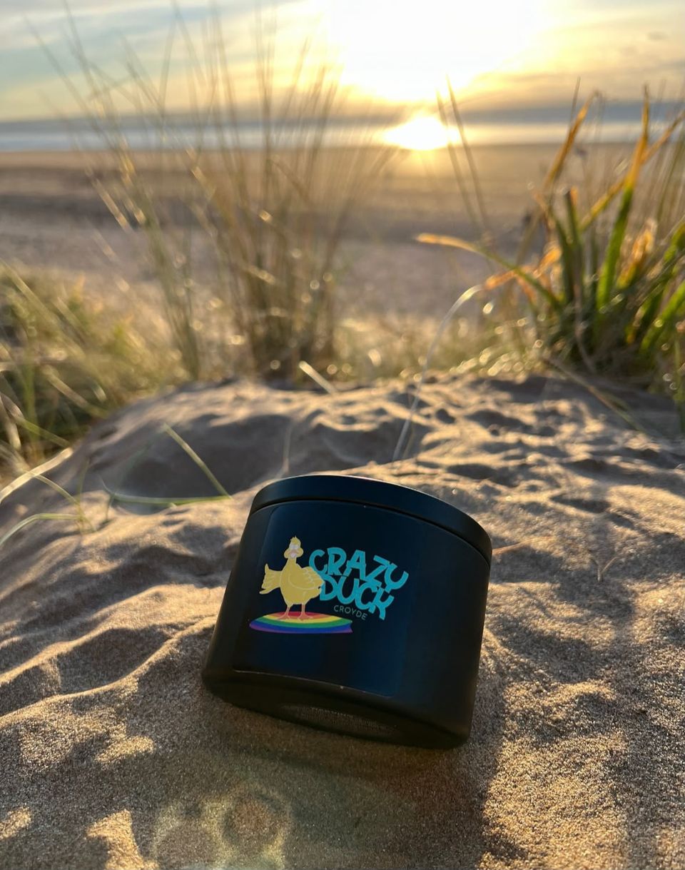 A Crazy Duck candle in its matte black tin, resting in the sand dunes at sunset