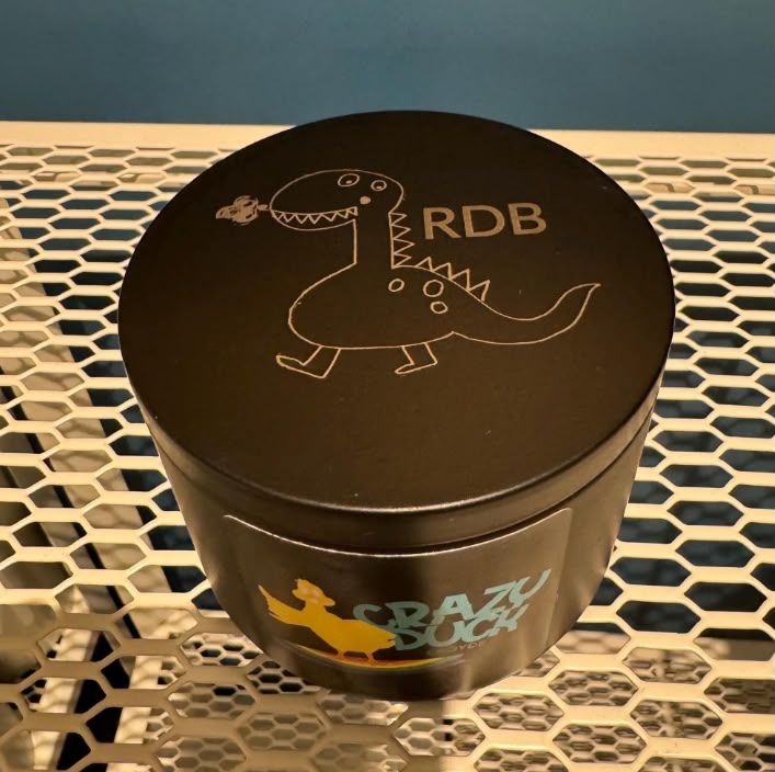An engraved candle lid showing a child's dinosaur drawing and the initials RDB